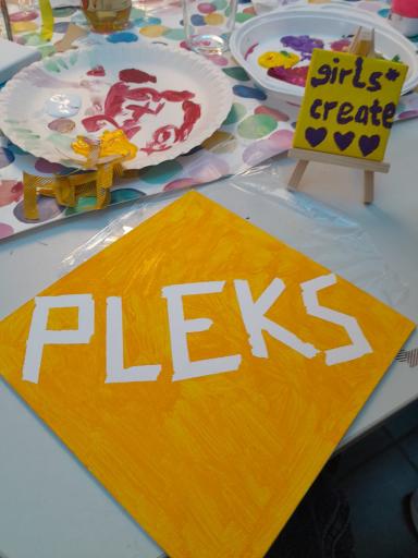 Plakat mit Logo von PLEKS liegt auf einem Tisch, daneben steht ein Schild, auf dem girls* create steht
