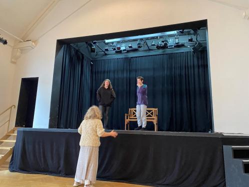 Zwei Mädchen* stehen auf der Bühne und haben Spaß. Eine Person steht vor der Bühne und spricht mit den Mädchen*.